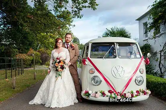 wedding car with burgundy ribbon and pink flowers, alongside a bride and groom
